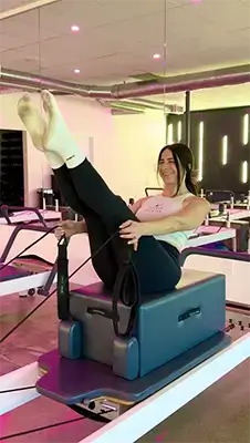 Member Juliana P. performing a challenging Pilates exercise on the Wunda Chair, lifting her legs toward the ceiling with excellent form in the studio.