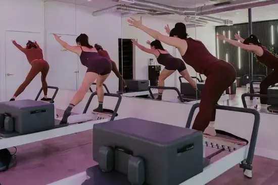 Group of women performing a deep balance and extension exercise on Pilates reformers in the brightly lit, modern Avenir Burbank studio with mirrors and pink accent lighting.